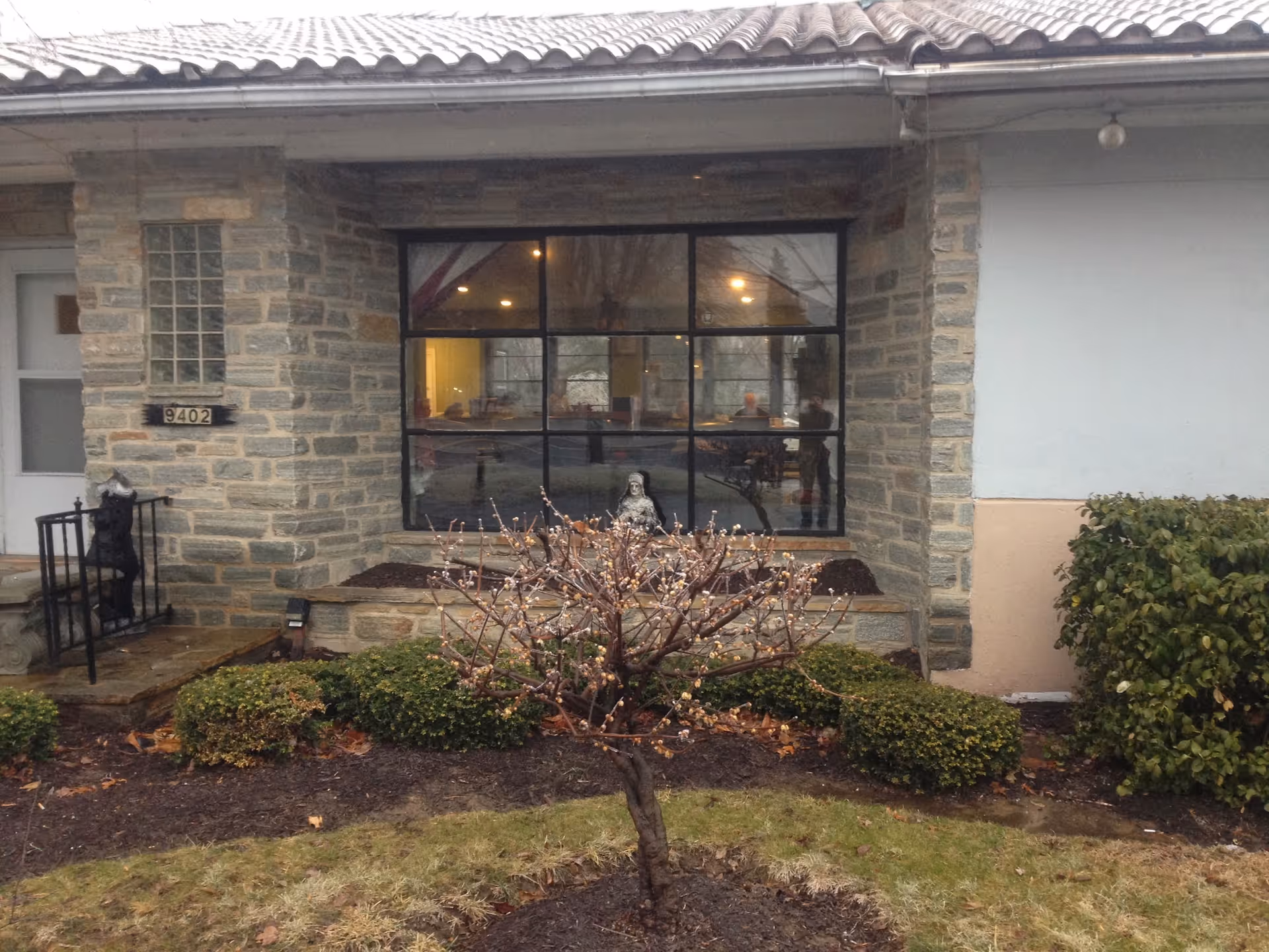 Stone-front building entrance with a large multi-pane window, house number 9402, and a small landscaped area with shrubs and a bare ornamental tree in front.