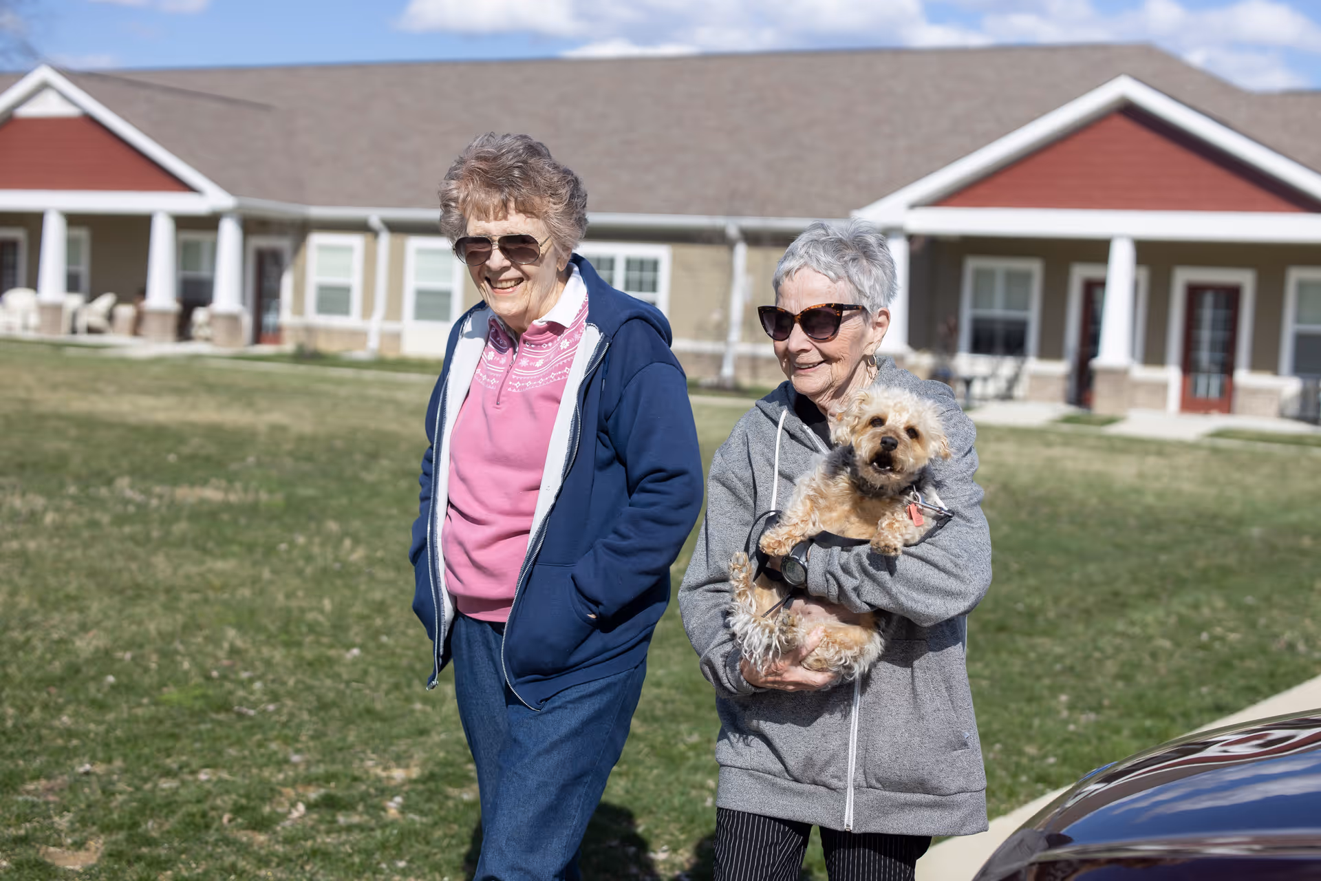 Two elderly women wearing sunglasses are outdoors on a grassy area in front of a single-story building with red and beige exterior walls. One woman is wearing a pink shirt and blue jacket, while the other is holding a small dog and wearing a gray hoodie. Both women are smiling and appear to be enjoying a sunny day.