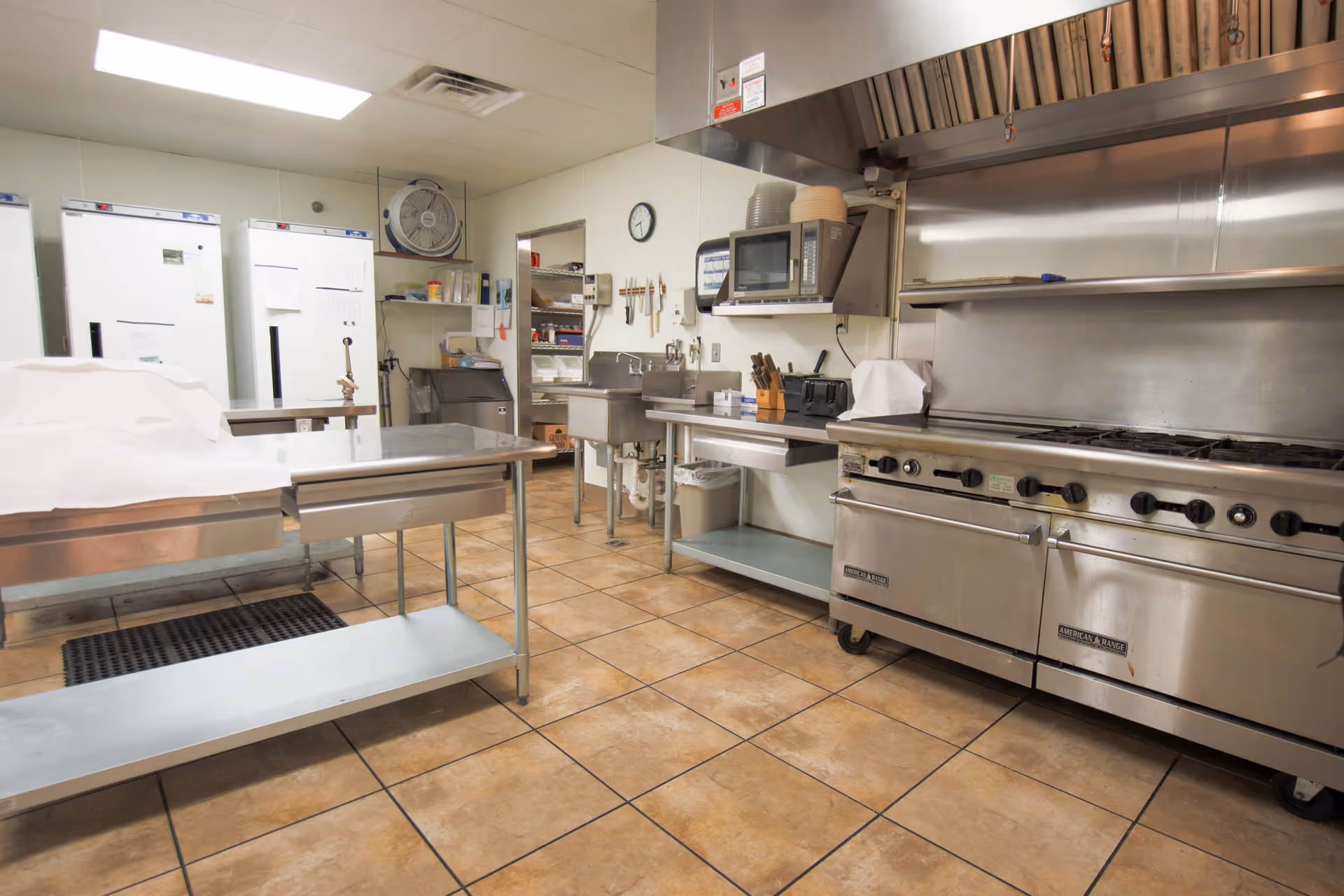 A clean commercial kitchen with stainless steel appliances and countertops, including a large stove with multiple burners, a microwave, sinks, refrigerators, and preparation tables. The floor is tiled and the walls are white.
