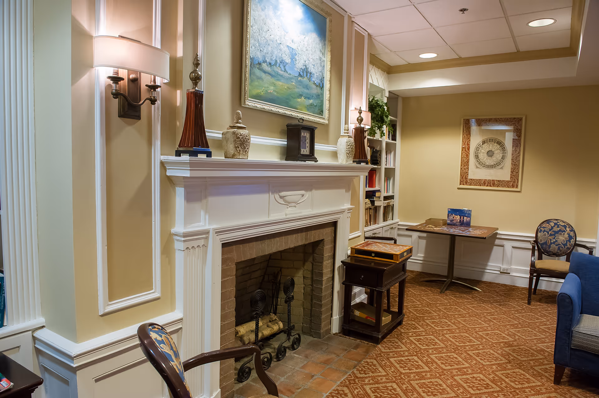 A cozy living room area featuring a white fireplace with decorative items on the mantel, including two lamps, vases, and a clock. Above the fireplace hangs a framed painting of a landscape. To the right, there is a small table with a puzzle and a chair with floral upholstery. The room has beige walls, built-in bookshelves, and a patterned carpet.