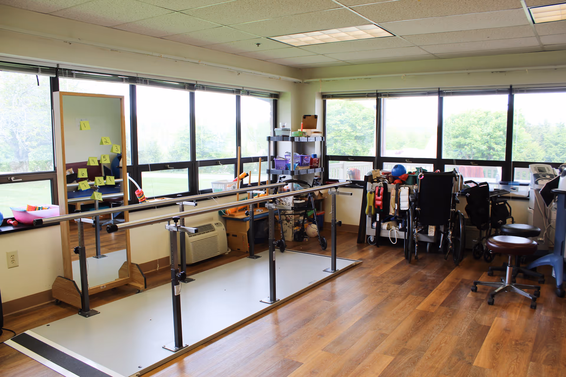 Bright therapy room with parallel walking bars, mobility aids and wheelchairs beside large windows overlooking greenery.