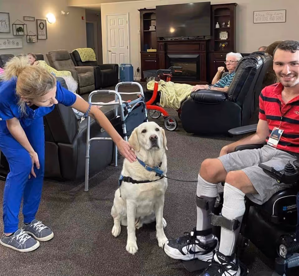 Residents and staff in a senior living lounge gathered around a sitting Labrador while a man in a wheelchair smiles.