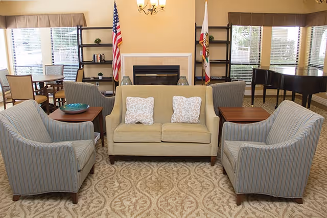 A cozy living room area with a beige loveseat adorned with two patterned pillows, flanked by two striped armchairs. Behind the loveseat is a fireplace with two flags on either side and shelves with decorative items. Large windows with blinds allow natural light to fill the room, and a grand piano is visible in the background.