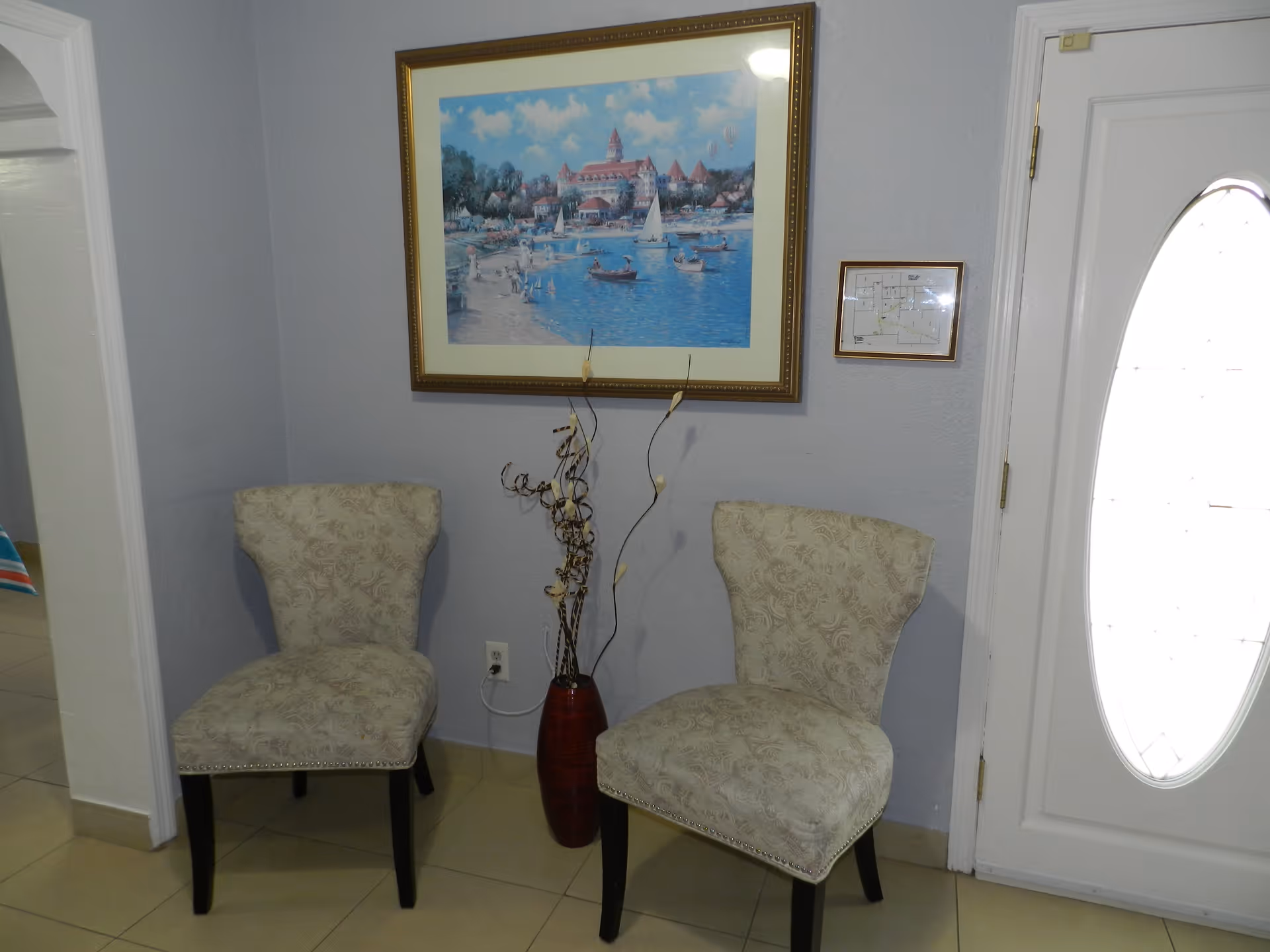 Two patterned beige chairs with dark wooden legs are placed against a light gray wall. Between the chairs is a tall red vase with decorative dried branches. Above the chairs hangs a framed painting of a lakeside scene with boats and a large building in the background. To the right is a white door with an oval glass window, and a small framed floor plan is mounted on the wall next to the door.