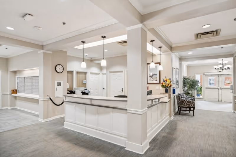 Bright and clean reception area in a senior living facility with a white front desk, beige walls, and carpeted floor. There are pendant lights hanging from the ceiling, a clock on the wall, mailboxes on the left side, and a seating area with a patterned chair and a plant near the entrance doors in the background.