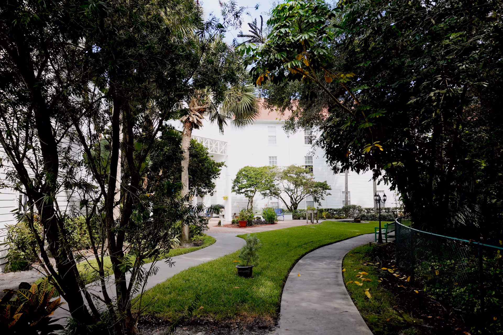Curved concrete walkway through a shaded courtyard with trees and a white multi-story building in the background.