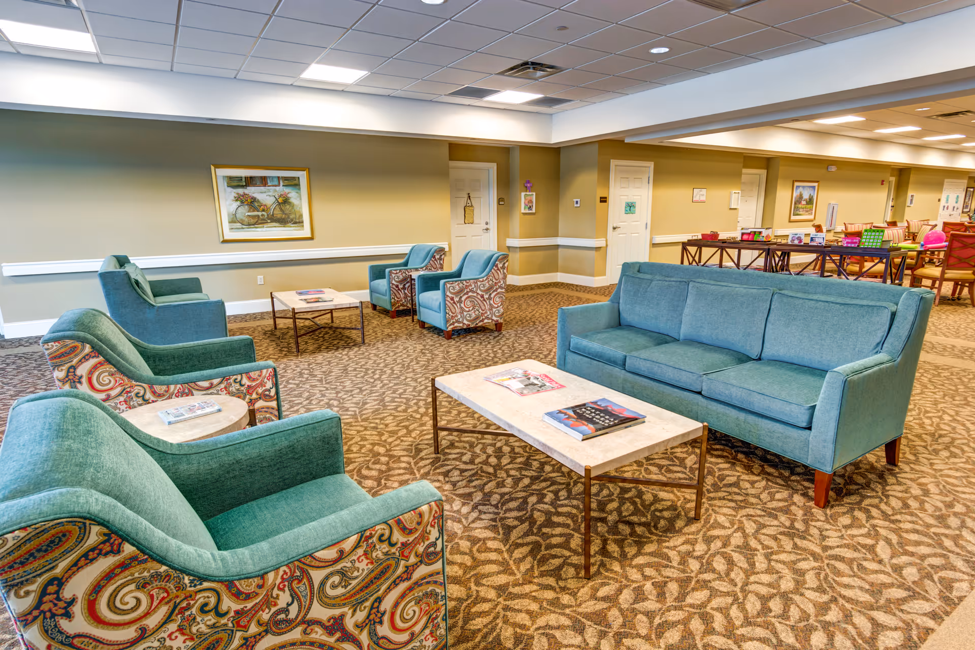 A spacious senior living facility common area with patterned carpet and beige walls. The room features a teal sofa, two teal armchairs with paisley-patterned sides, and two additional teal armchairs arranged around two rectangular coffee tables with magazines on top. In the background, there are tables and chairs set up for activities or dining, and framed artwork on the walls.