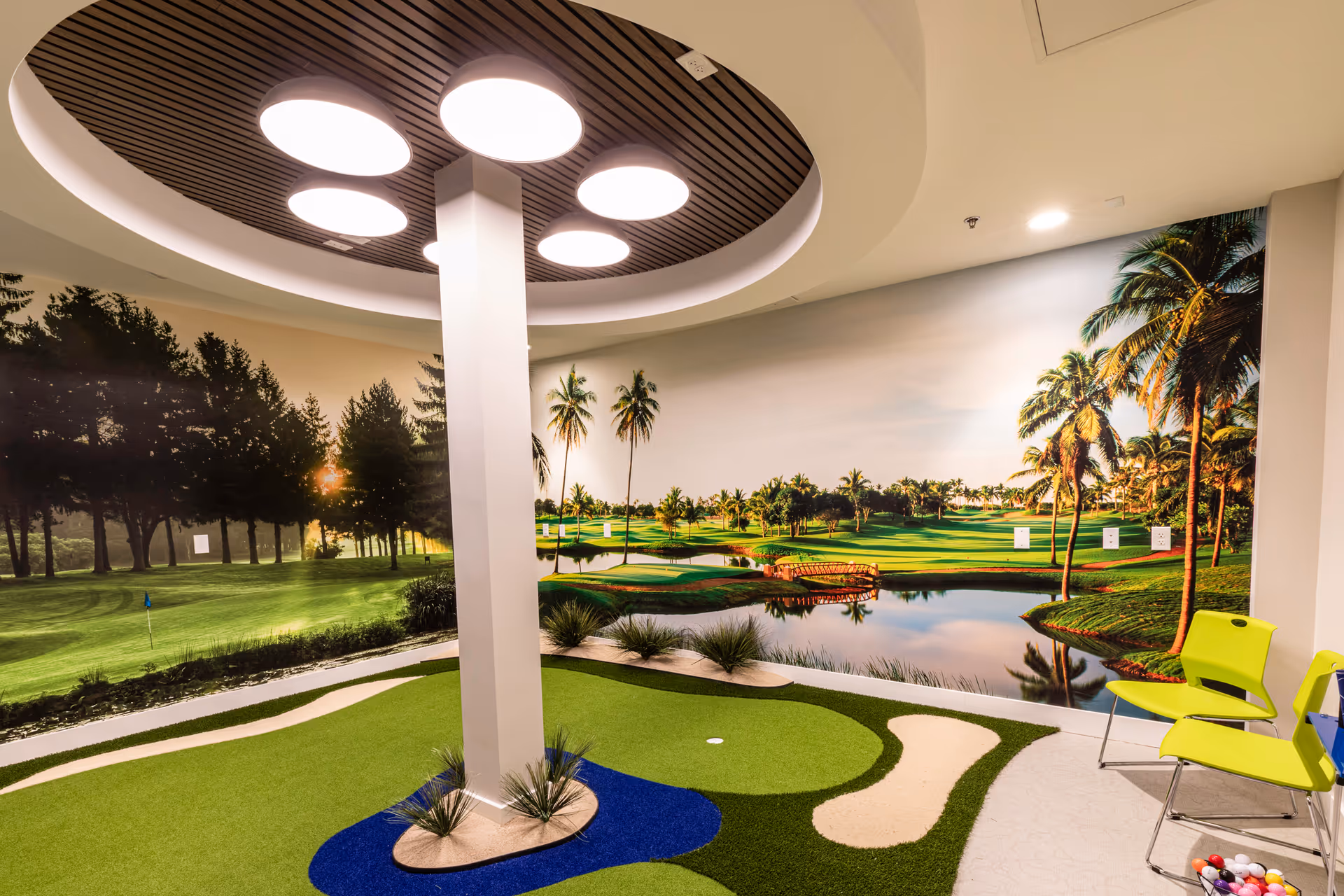 Indoor golf putting area with mural, circular ceiling lights, green turf, and yellow chairs.