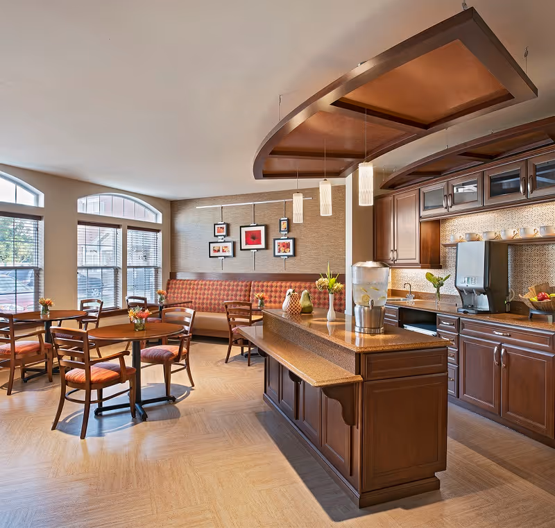 A bright dining area with wooden tables and chairs, a cushioned bench along the wall with framed artwork above it, large windows letting in natural light, and a kitchen counter with a water dispenser and decorative vases. The kitchen area has wooden cabinets and modern lighting fixtures hanging from the ceiling.