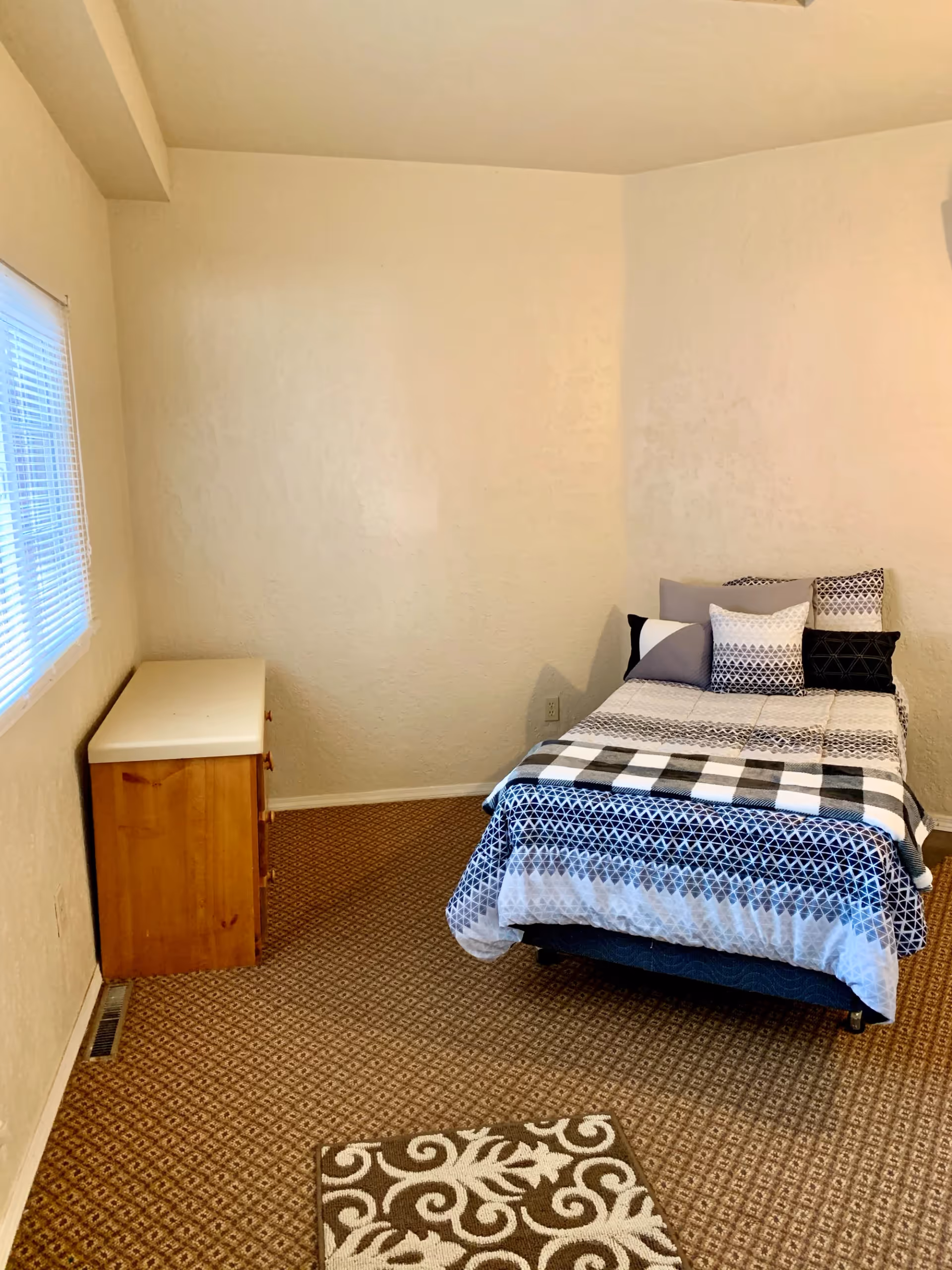 A small bedroom with beige walls and patterned carpet. There is a single bed with a geometric patterned bedspread and multiple pillows against the right wall. A wooden dresser with a white top is positioned against the left wall under a window with closed blinds. A small decorative rug is on the floor near the foot of the bed.