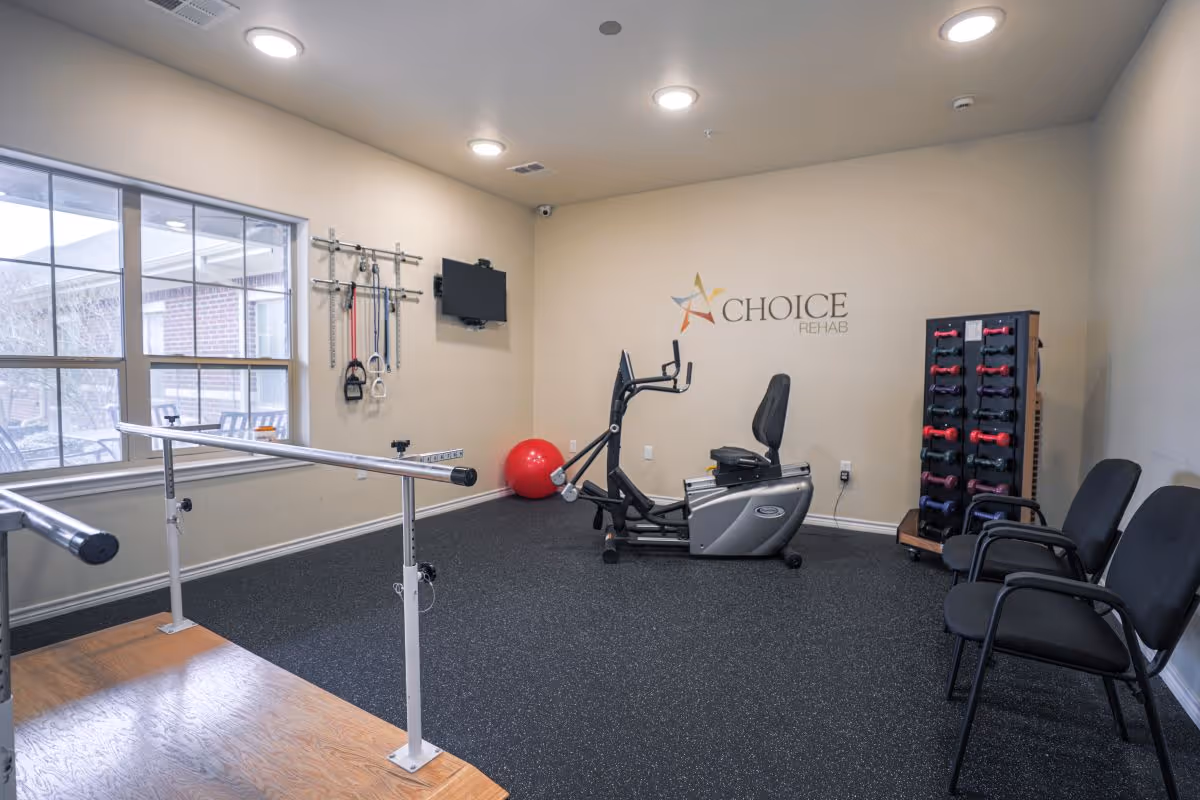 A small rehabilitation exercise room with a recumbent exercise bike, parallel bars for walking support, a rack of colorful dumbbells, resistance bands hanging on the wall, a red exercise ball, three black chairs, and a wall-mounted TV. The room has beige walls and a window showing an outdoor patio area. The wall features the logo 'CHOICE REHAB'.