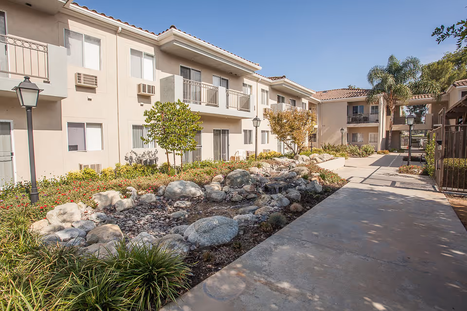 Outdoor courtyard area of a senior living facility with a paved walkway, landscaped garden featuring rocks, plants, and small trees, and a two-story beige building with balconies and windows under a clear blue sky.