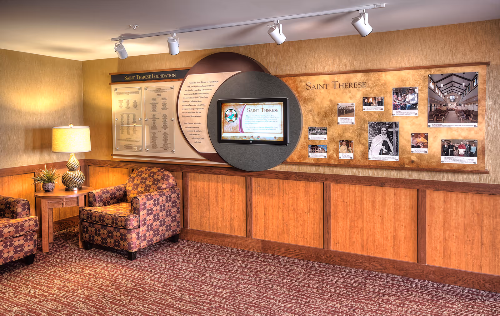 A cozy senior living lounge with patterned armchairs, a side table and lamp, and a wall display labeled "Saint Therese" with a central digital screen.