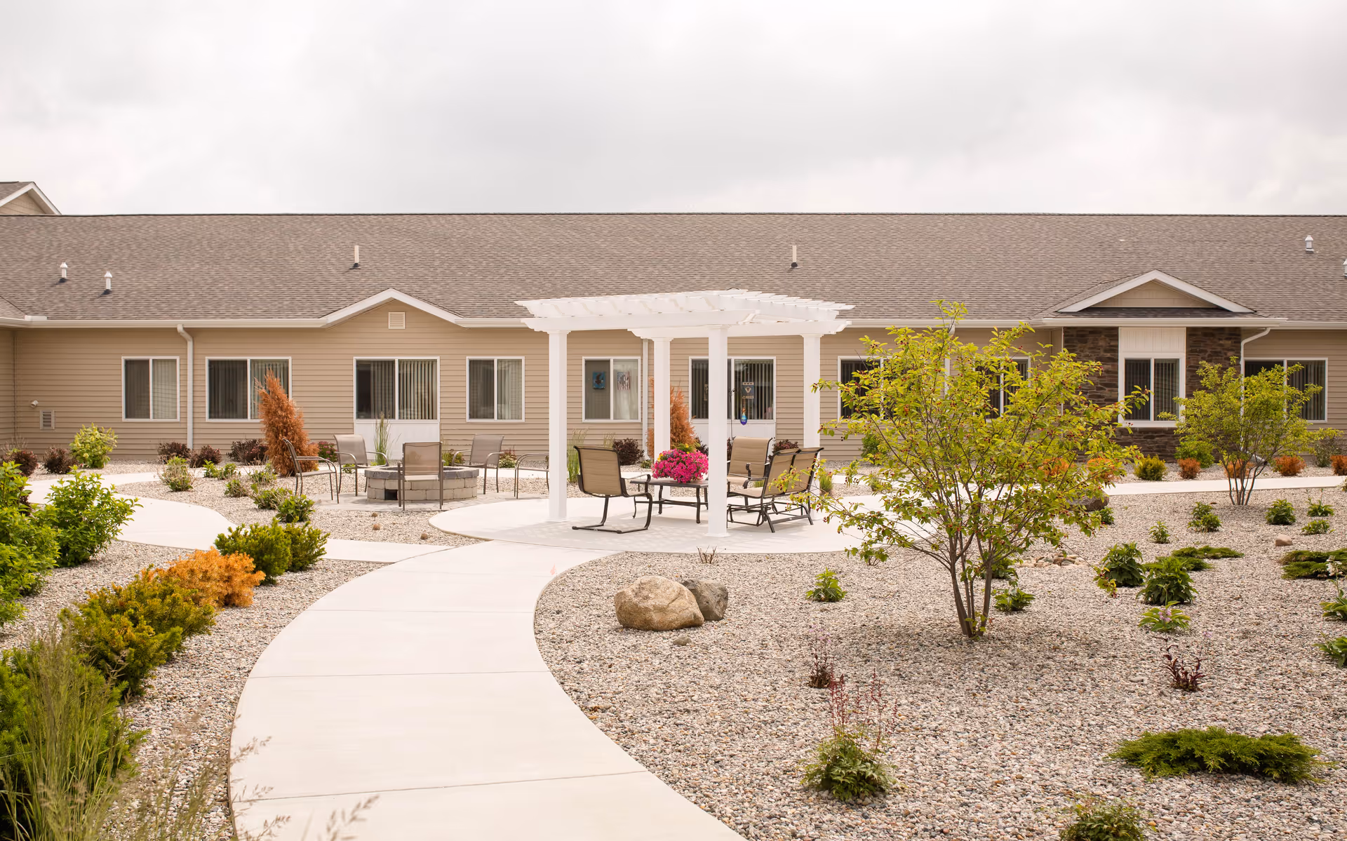 Courtyard with a winding paved path, a white pergola and outdoor seating in front of a single-story building.