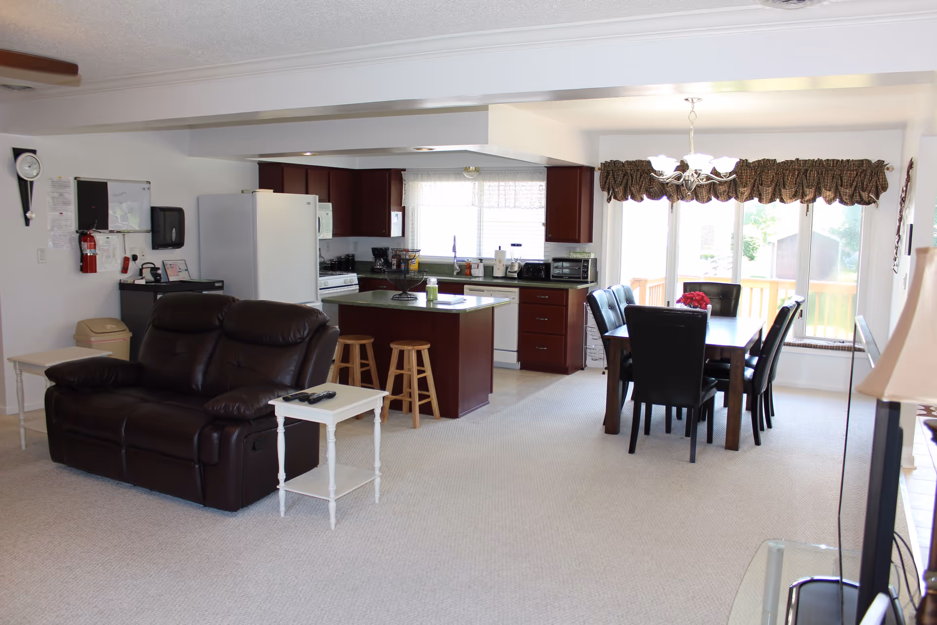 Open interior space featuring a living room with a dark brown leather loveseat and a small white side table with remote controls, adjacent to a kitchen with dark wood cabinets, a white refrigerator, stove, microwave, and island with two wooden stools. To the right, there is a dining area with a dark wood table, six black leather chairs, a chandelier overhead, and large windows with patterned valances letting in natural light.