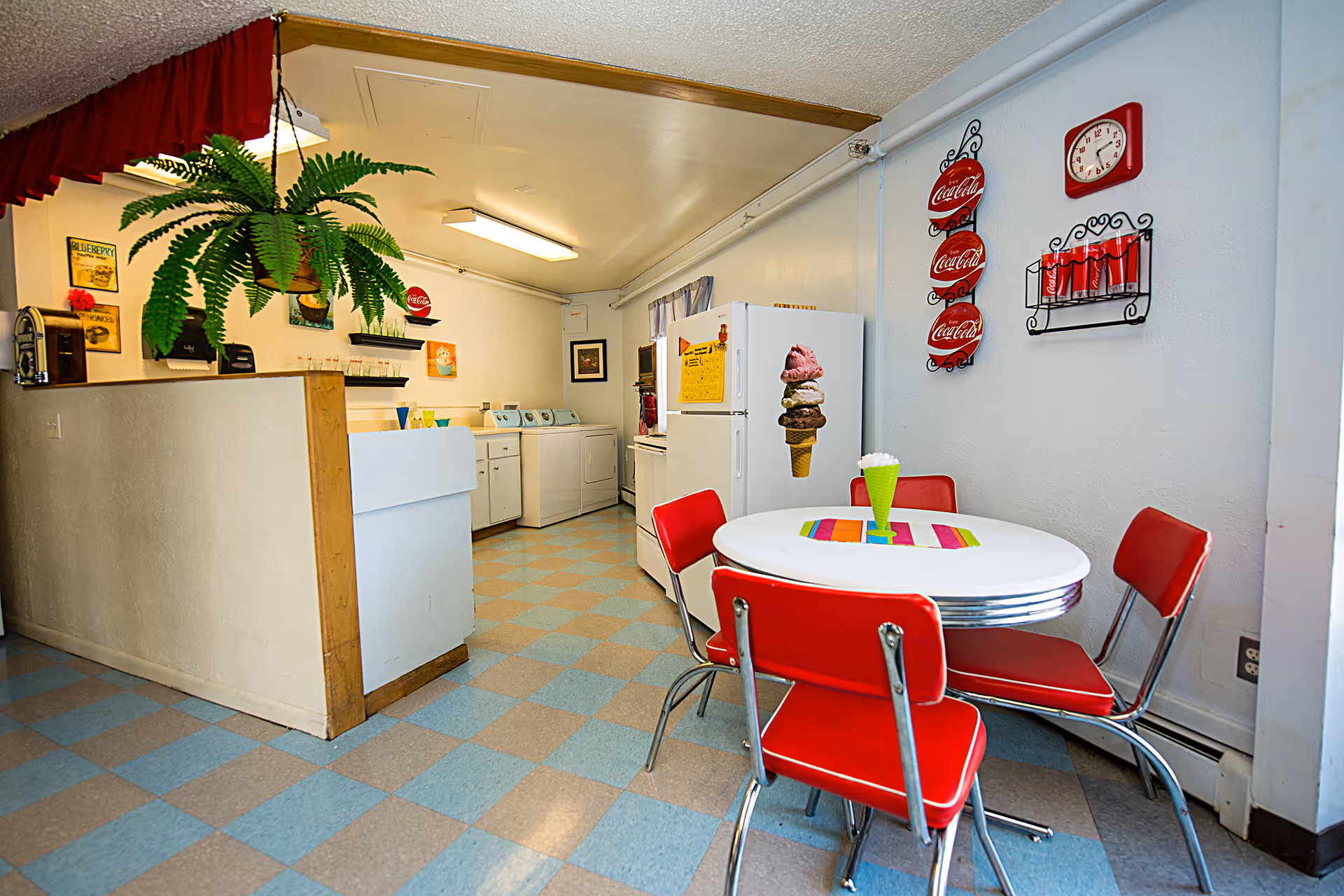 A retro-style kitchen and dining area with a round white table surrounded by four red chairs. The walls are decorated with Coca-Cola themed items including plates, a clock, and a rack holding Coca-Cola cans. The kitchen area has a white refrigerator with an ice cream cone magnet, a stove, and a laundry area with a washer and dryer. A green hanging plant is visible above a half wall, and the floor is covered with blue and gray checkered tiles.