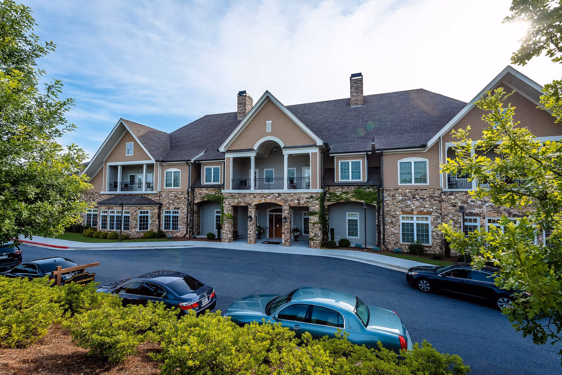 Front exterior view of a large two-story senior living facility building with stone and beige siding, multiple windows, balconies, and a covered entrance. Several cars are parked in front, and green trees and bushes surround the area under a partly cloudy sky.