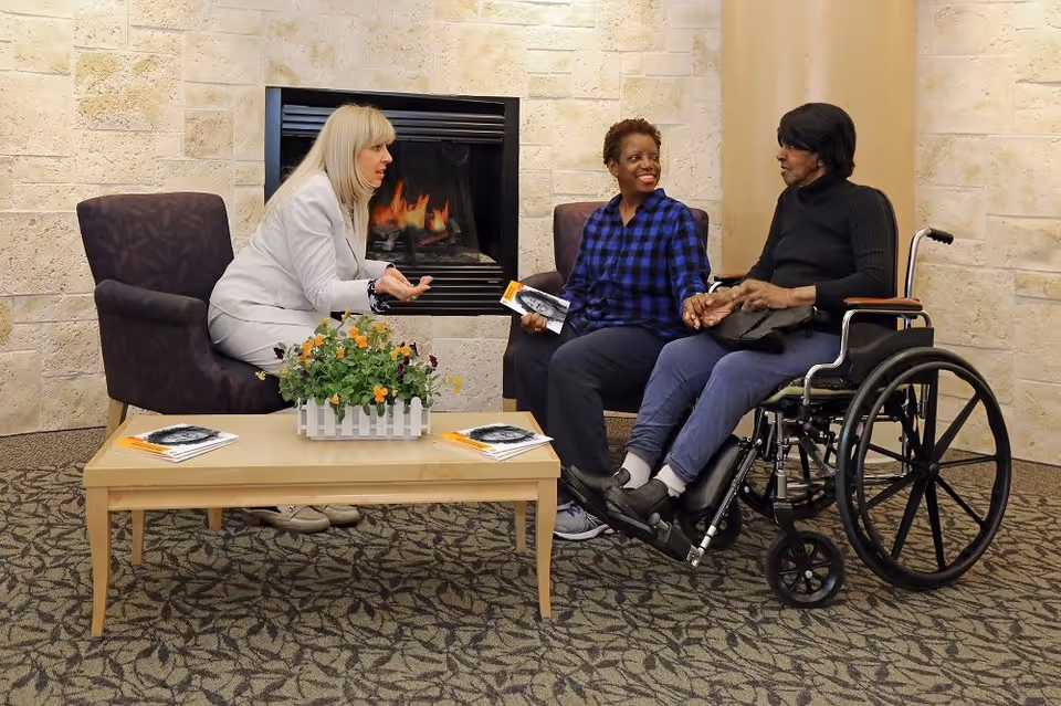 Three women sitting and talking in a cozy room with a fireplace. One woman is seated in a wheelchair, another is sitting on a chair holding her hand, and the third woman is sitting across from them on another chair. A coffee table with a flower arrangement and brochures is in front of them.