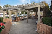 Outdoor seating area with a wooden pergola overhead, surrounded by brick walls and bushes. Several chairs and tables are arranged under the pergola, with parked cars and trees visible in the background.