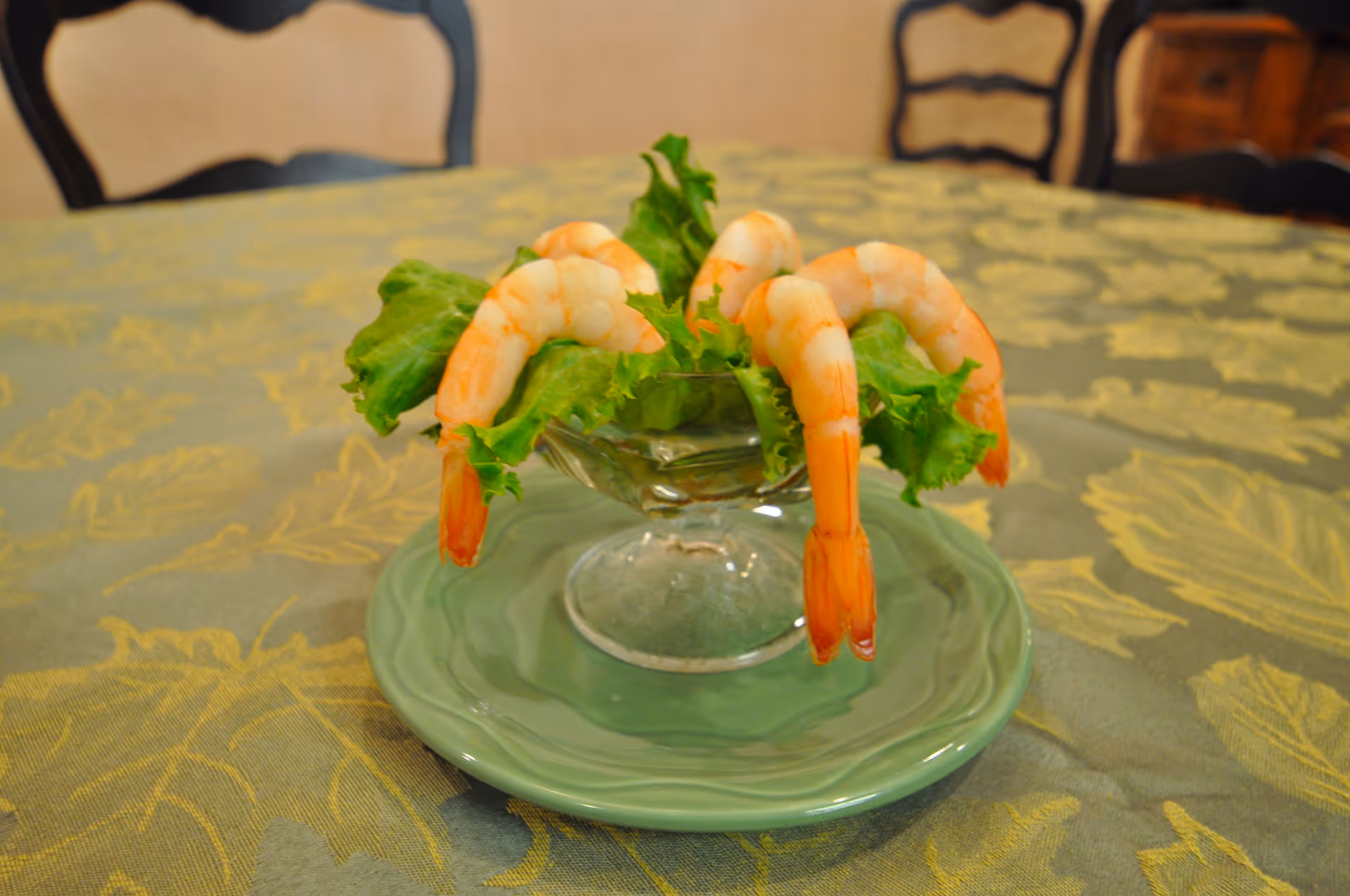 A glass dish with several cooked shrimp arranged around the rim, resting on green lettuce leaves. The dish is placed on a green plate on a table covered with a patterned tablecloth. In the background, there are wooden chairs and a piece of furniture.