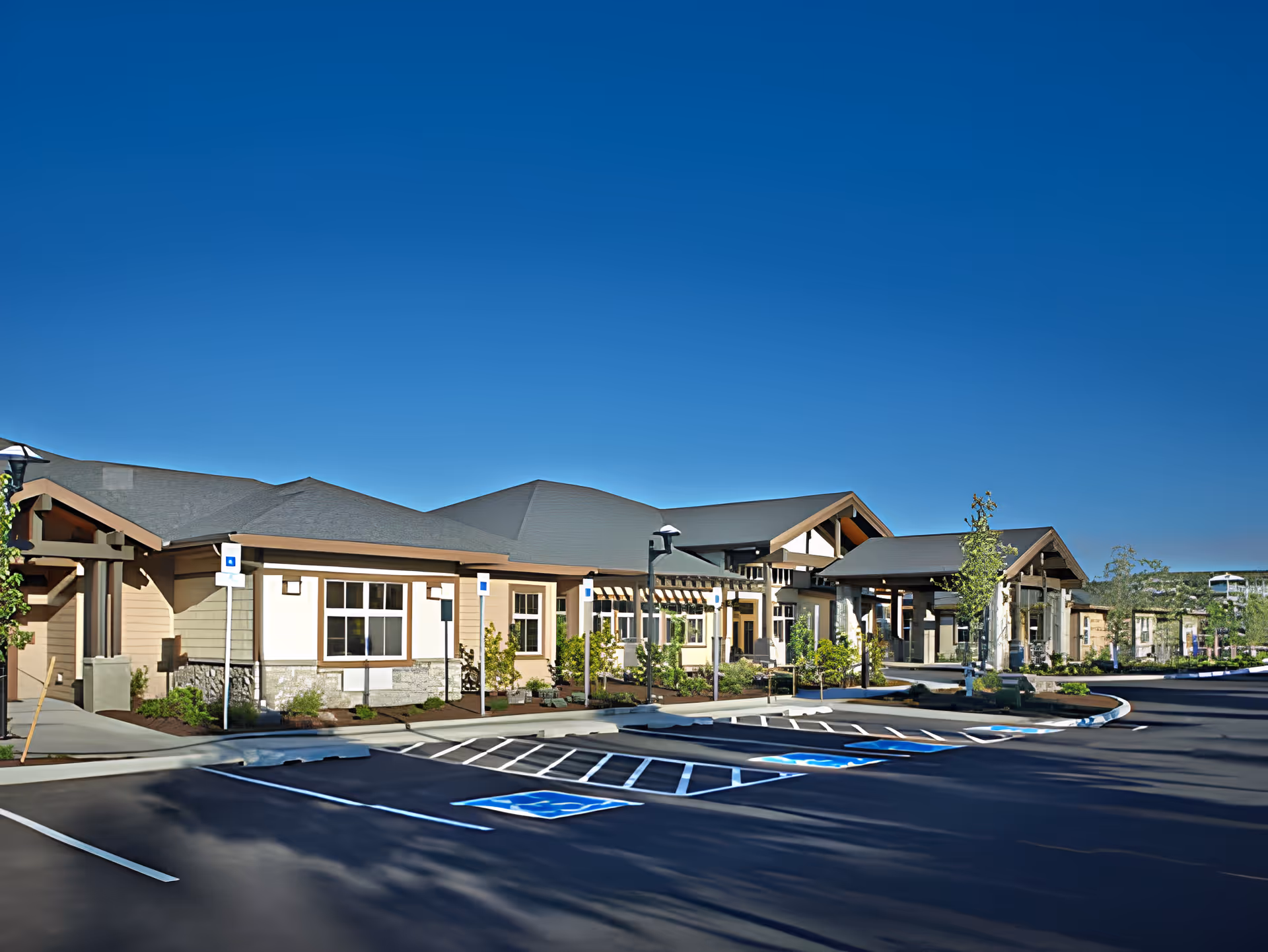 Exterior view of Bend Transitional Care facility showing a single-story building with a covered entrance, multiple windows, landscaped greenery, and a parking lot with marked handicap spaces under a clear blue sky.