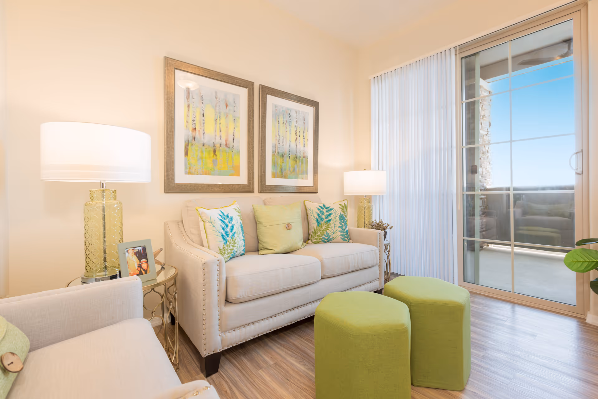 A bright and cozy living room with a beige sofa adorned with green and blue leaf-patterned cushions, two green ottomans, a beige armchair, two table lamps with yellow glass bases, framed abstract artwork on the wall, and a sliding glass door leading to an outdoor balcony.