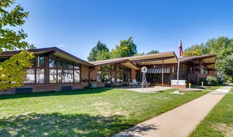 Exterior view of a single-story brick building with large windows and a slanted roof, surrounded by green grass and trees under a clear blue sky. A sidewalk leads up to the entrance, where an American flag and a small windmill decoration are visible.