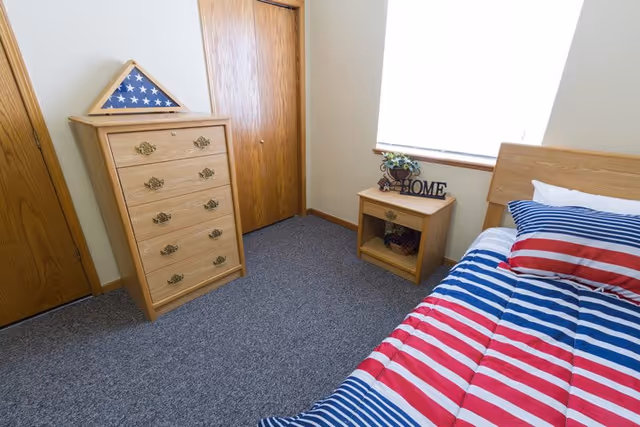 A small bedroom with a bed covered in a red, white, and blue striped comforter and matching pillow. Next to the bed is a wooden nightstand with a decorative plant and a 'HOME' sign. Across from the bed is a wooden dresser with a folded American flag in a triangular display case on top. The room has carpeted flooring and wooden doors.