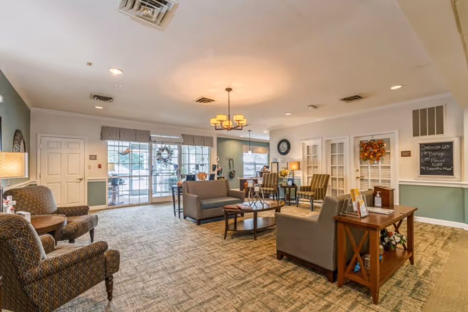 A spacious and well-lit senior living facility common area with comfortable seating including armchairs and sofas arranged around coffee tables. The room features carpeted flooring, a chandelier, large windows with valances, and a door decorated with a fall wreath. There is a small table with informational brochures and hand sanitizer, and a chalkboard on the wall listing activities such as Bingo and Movie Night.