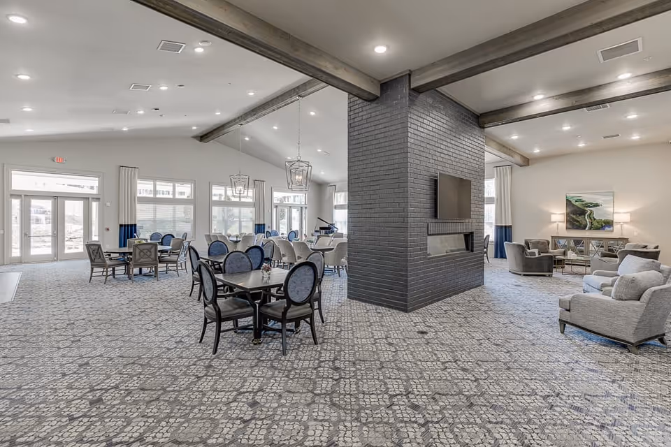 Spacious assisted living facility common area with patterned carpet, multiple dining tables with chairs, a large central black brick fireplace with a mounted TV, and a seating area with gray armchairs and a sofa. Large windows and glass doors allow natural light to fill the room.