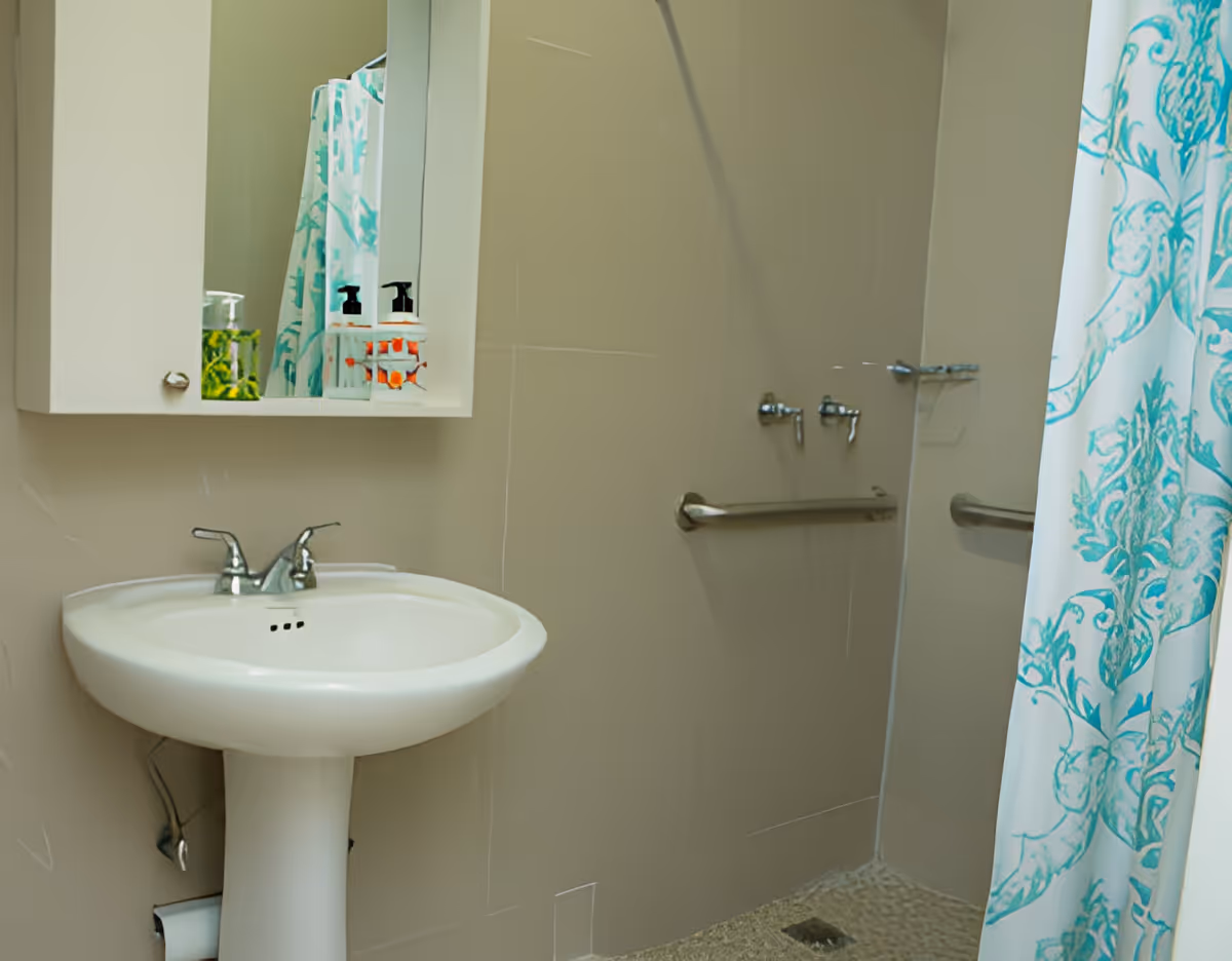 A bathroom with a white pedestal sink, a wall-mounted mirror cabinet with soap dispensers, a shower area with grab bars, and a shower curtain with a blue and white pattern.