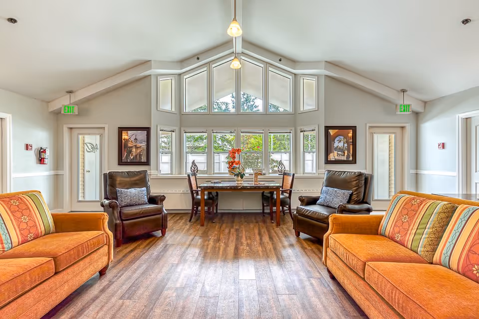 A bright and spacious living room area with two orange sofas featuring patterned cushions, two dark brown leather armchairs with pillows, and a wooden table with four chairs near large windows letting in natural light. The room has high vaulted ceilings with hanging pendant lights and wood flooring.