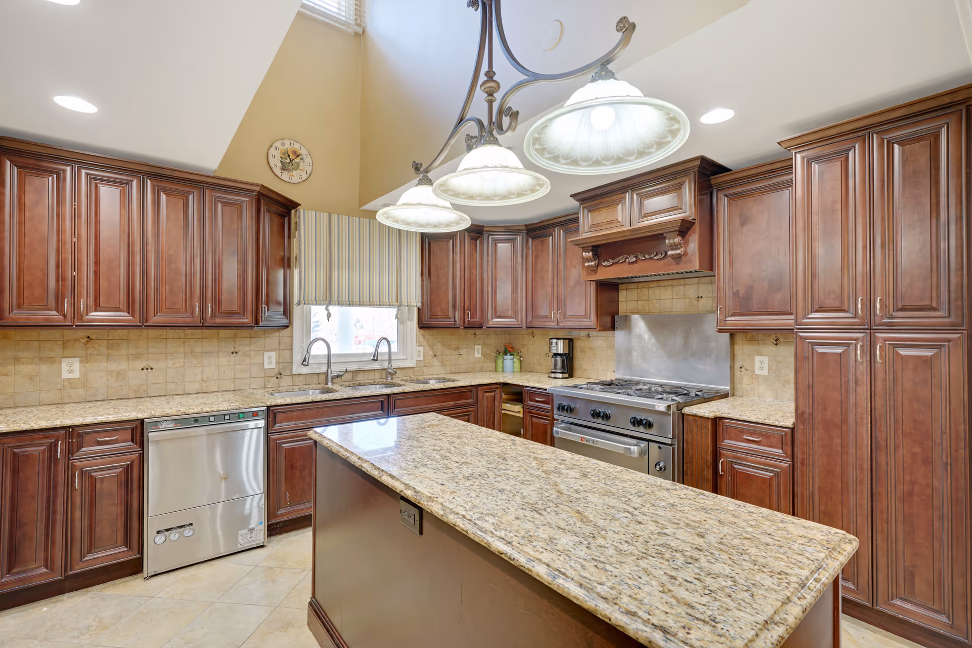 A spacious kitchen with dark wooden cabinets, granite countertops, a central island, stainless steel dishwasher and stove, a double sink under a window with a striped valance, and three hanging pendant lights above the island.