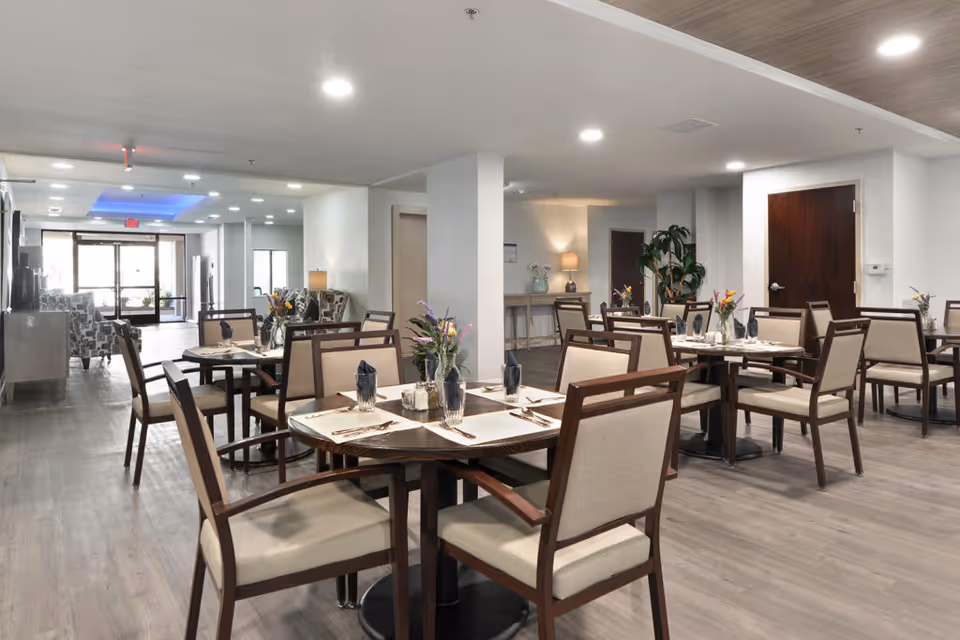A dining area in a senior living facility with multiple round tables set with placemats, napkins, silverware, and small flower arrangements. The room has wooden chairs with beige cushions, light-colored walls, and a wood floor. In the background, there is a seating area with patterned armchairs near large windows and a door leading outside.