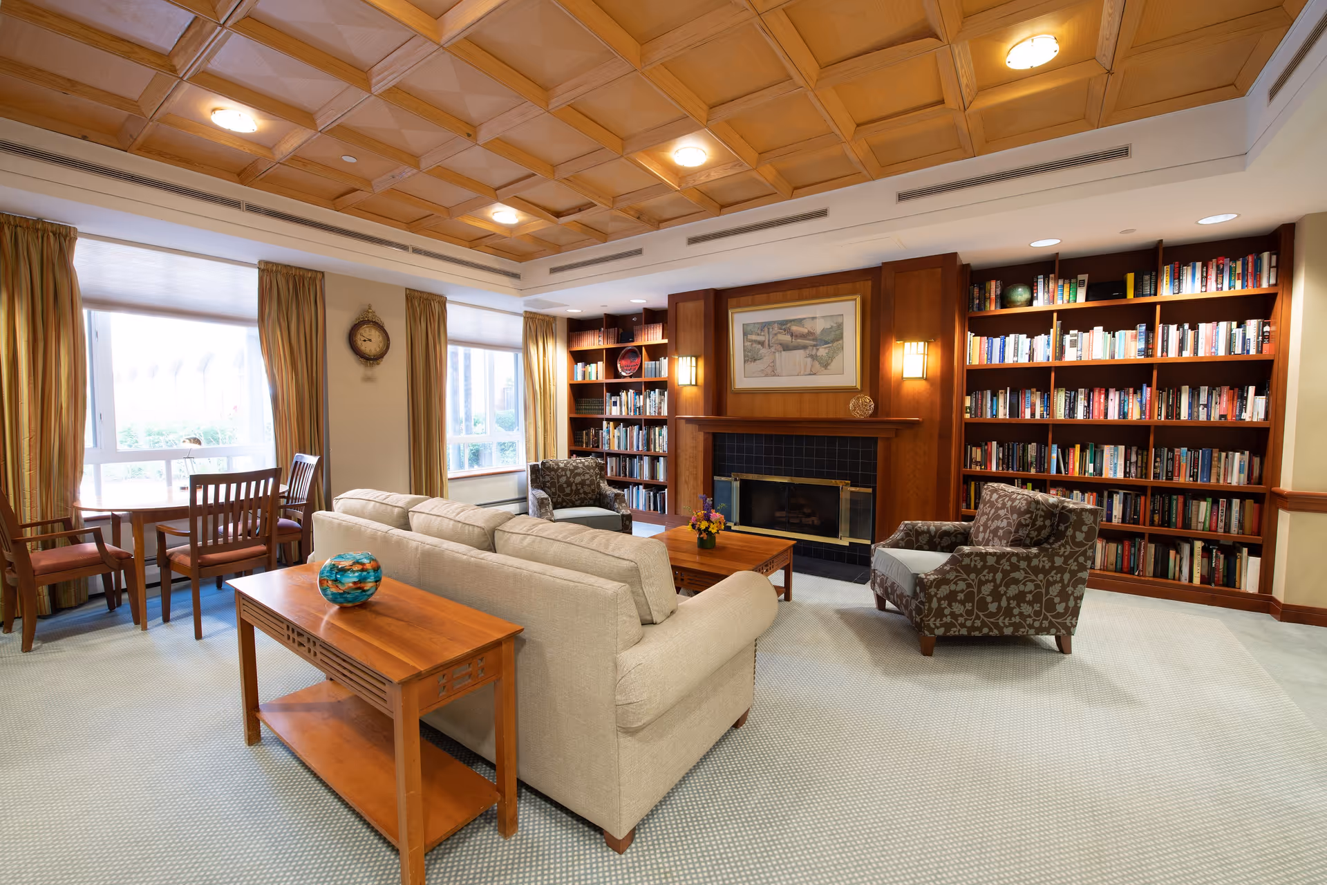 A cozy living room area in an assisted living facility featuring a beige sofa, two patterned armchairs, a wooden coffee table with a small flower arrangement, and a wooden side table with a decorative glass bowl. The room has built-in wooden bookshelves filled with books, a fireplace with a framed painting above it, large windows with golden curtains, and a wooden coffered ceiling with recessed lighting.