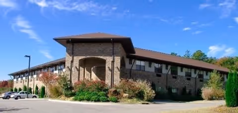 Exterior view of a two-story senior living facility building with a stone facade and a brown roof under a blue sky with some clouds. There are shrubs and greenery around the building and a parking area with a few cars visible.