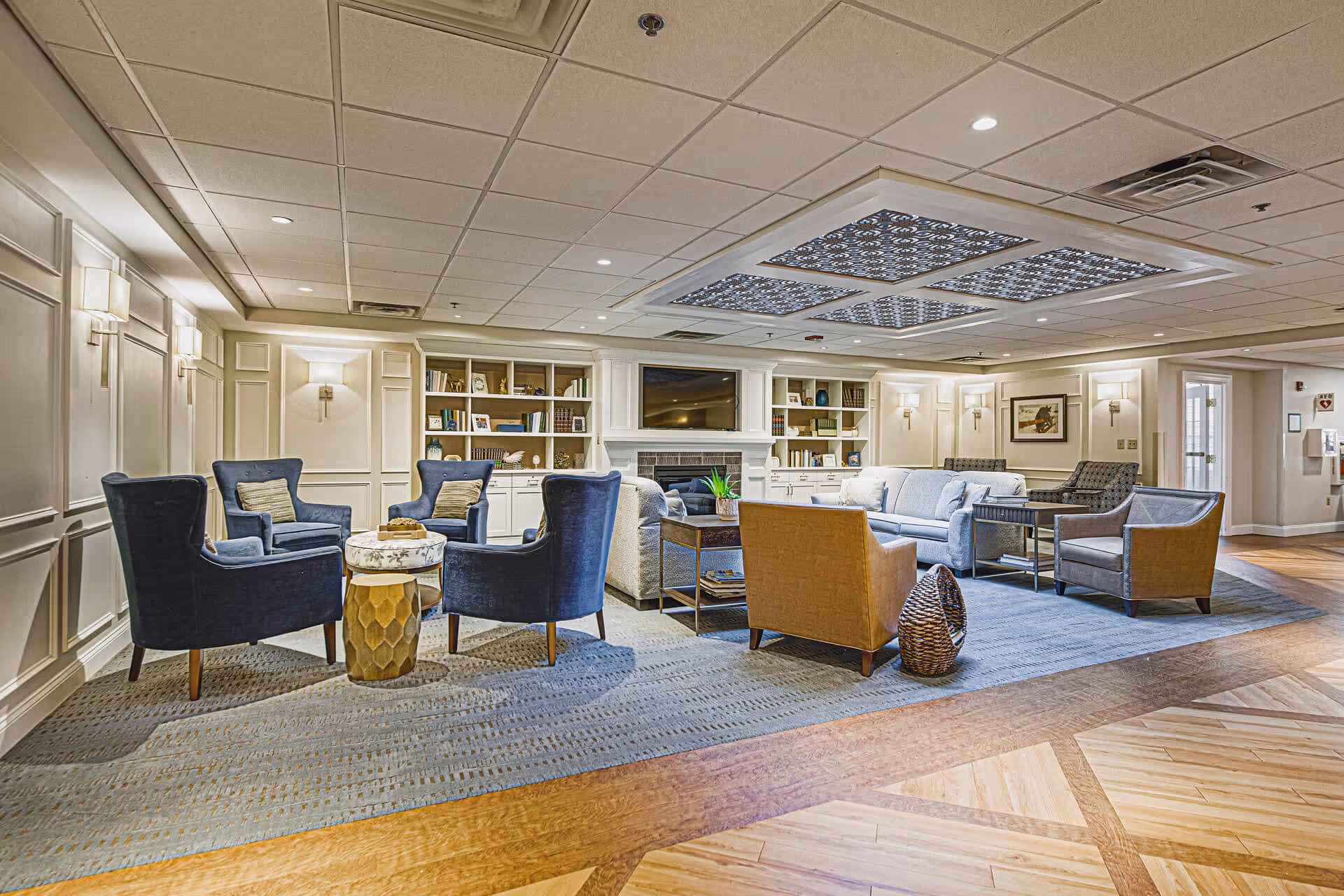 A spacious and well-lit living room area with multiple seating options including blue armchairs, a beige armchair, and a light blue sofa arranged around a central coffee table. The room features built-in white bookshelves filled with books and decorative items, a fireplace with a mounted TV above it, and elegant wall sconces providing soft lighting. The ceiling has decorative panels with intricate patterns, and the floor is a combination of wood and a large area rug.