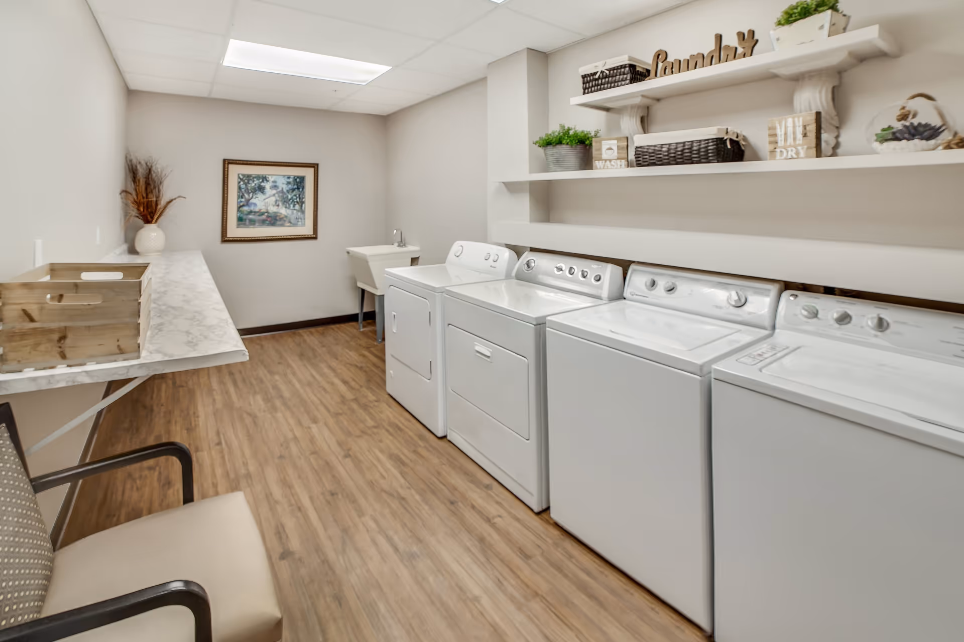 Laundry room with three white washing machines and dryers lined up against the right wall, a utility sink in the back corner, wooden shelves above holding baskets, plants, and decorative signs including one that says 'Laundry'. There is a chair and a folding table with a wooden crate on the left side, wood-look flooring, and a framed picture on the back wall.