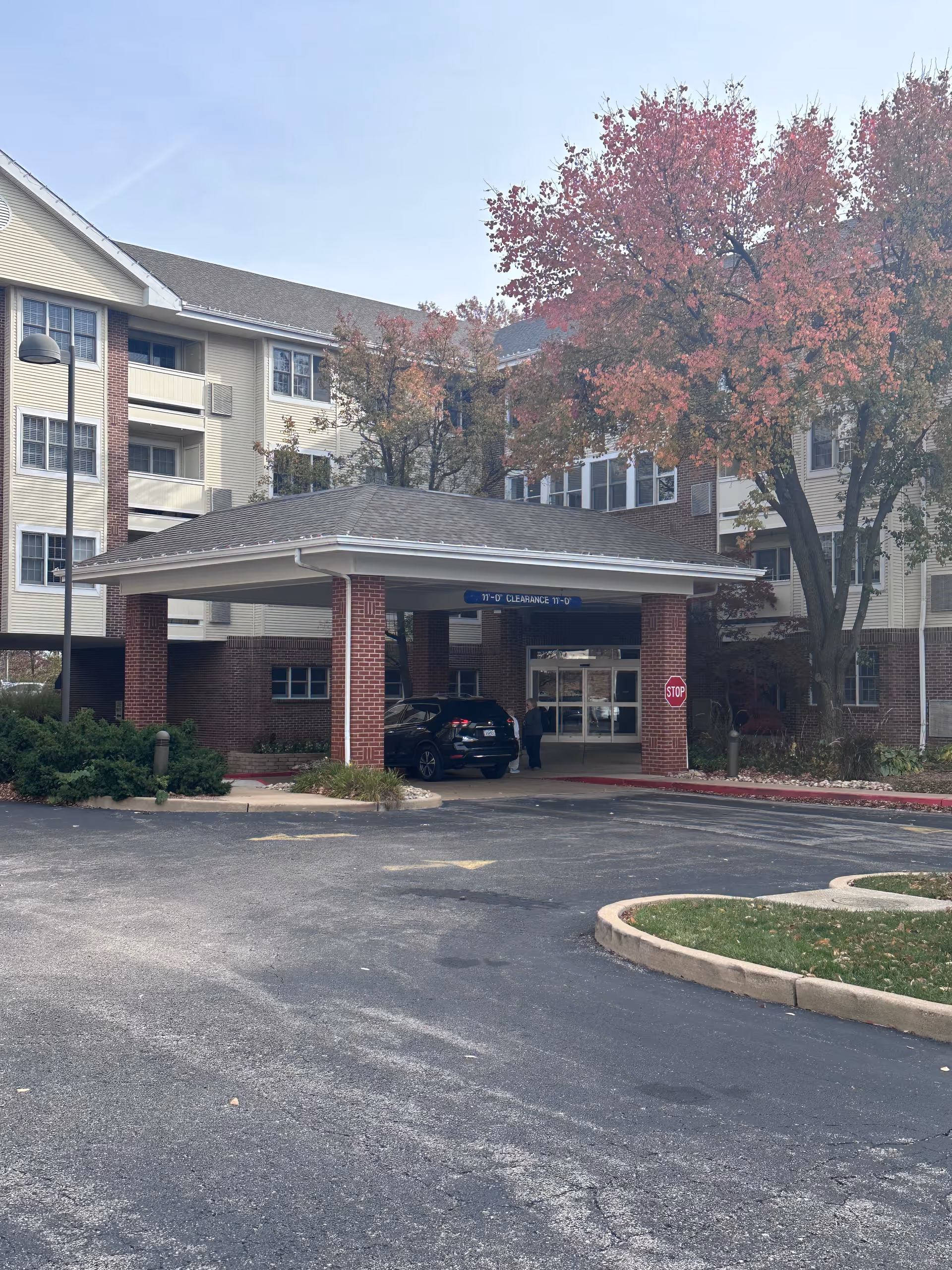 Entrance of a senior living facility named Bethesda Gardens with a covered drop-off area supported by brick pillars. A black SUV is parked under the canopy, and a person is standing near the vehicle. The building is multi-story with beige siding and brick accents. Trees with autumn-colored leaves are visible near the entrance, and a stop sign is posted by the driveway.