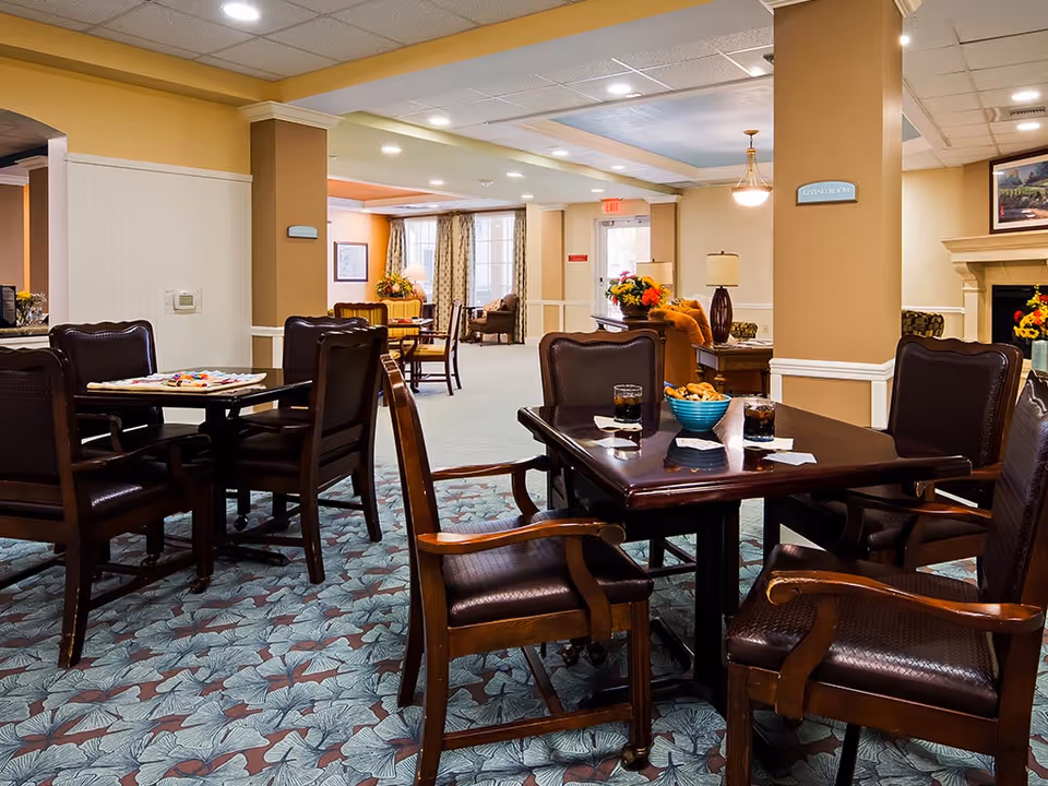 A cozy living room area in The Windsor Of Ocala featuring dark wooden tables and chairs with cushioned seats. On the tables are glasses of soda and a bowl of snacks. The room has patterned carpet, beige walls with white trim, and a fireplace in the background. There are also floral arrangements and lamps adding to the warm ambiance.
