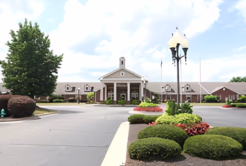 Front exterior view of Five Star Residences of Clearwater, showing a large building with a central entrance featuring columns, surrounded by landscaped bushes, flowers, a tree, and a lamp post under a partly cloudy sky.