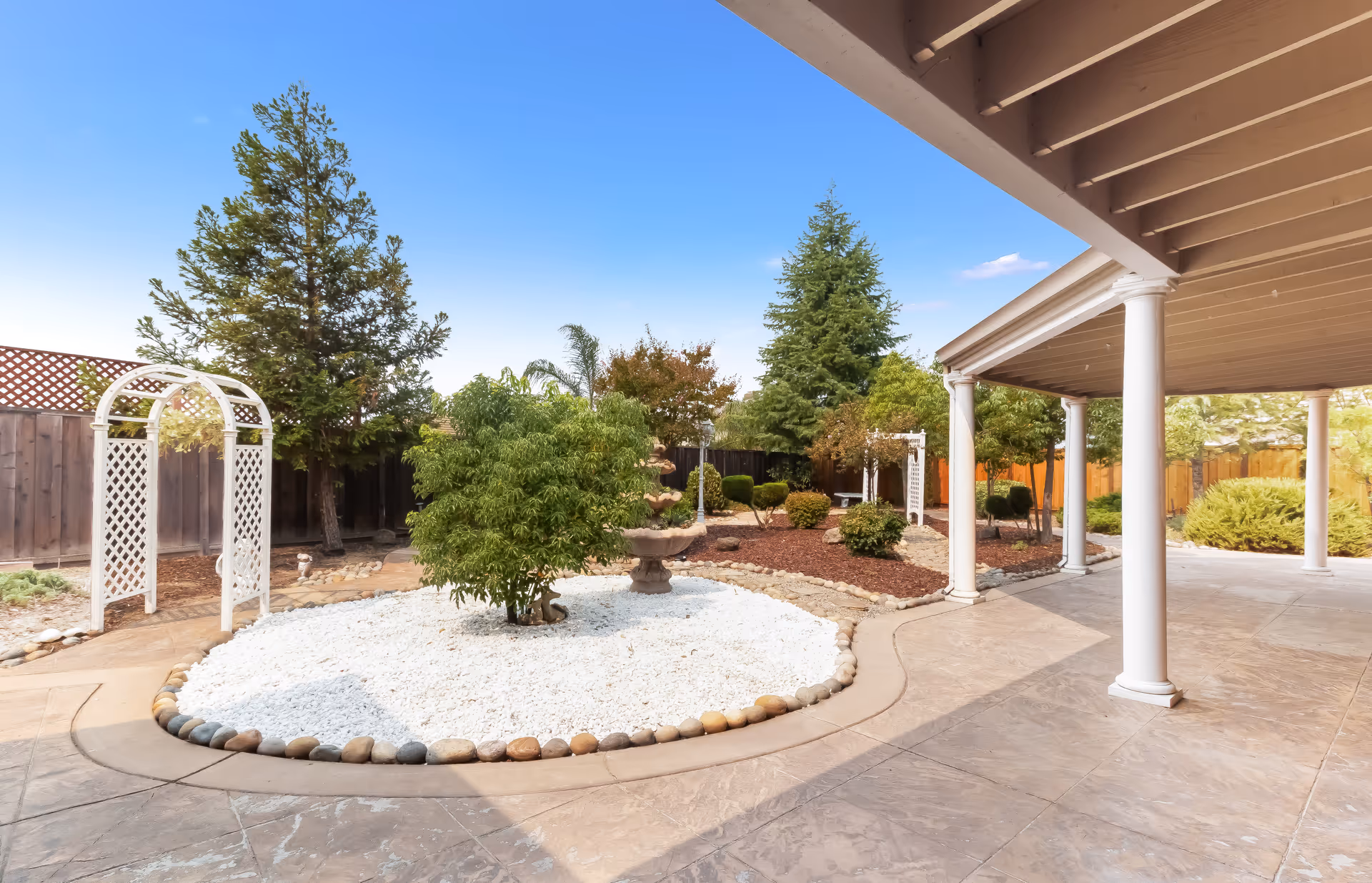A landscaped outdoor garden area with a white gravel bed surrounded by smooth stones, a small bush, and a multi-tiered stone fountain. The garden is enclosed by a wooden fence and features several trees and shrubs. There is a covered patio with white columns and a concrete floor on the right side of the image under a clear blue sky.