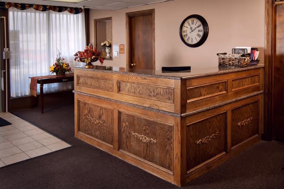 Wooden reception desk in a lobby with floral arrangements, a wall clock, and curtained windows.
