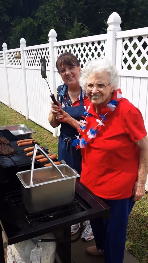 An elderly woman wearing a red shirt and a red, white, and blue flower lei is smiling while holding a spatula at a grill. A younger woman stands behind her, also smiling and wearing a blue apron. They are outdoors next to a white fence with grass and trees in the background. Hot dogs and burgers are cooking on the grill.