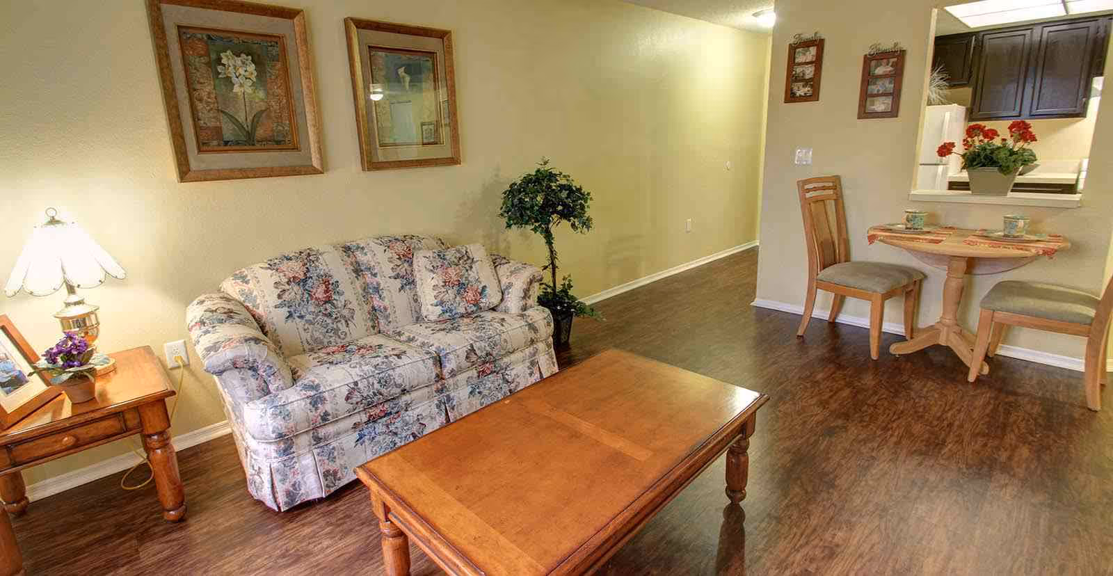 Cozy living area with a floral sofa, wooden coffee and side tables, a lamp, a small round dining table with two chairs, and a kitchen pass-through.