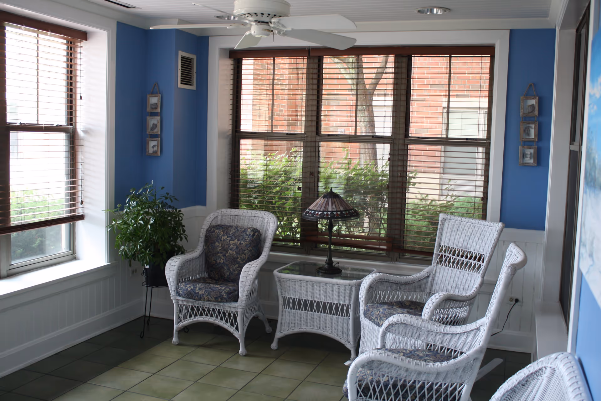 A cozy sitting area with white wicker chairs and a matching wicker table with a decorative lamp on it. The room has blue walls, large windows with wooden blinds, a ceiling fan, and a potted plant in the corner.