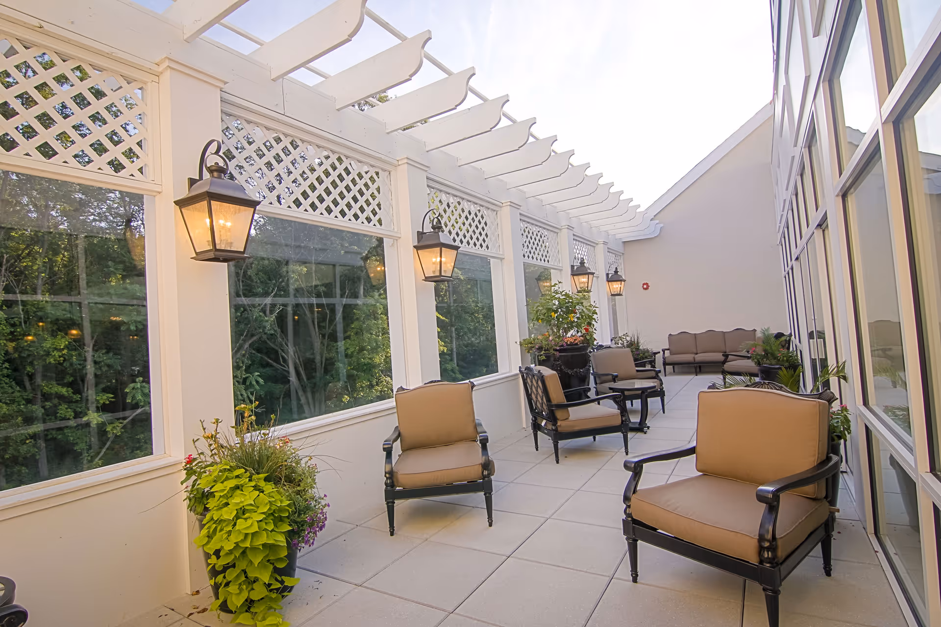 A bright outdoor patio area at Brandywine Mahwah by Monarch featuring cushioned armchairs and a sofa arranged along the sides. The space is enclosed with large windows and white lattice panels above, with black lantern-style wall lights mounted on the pillars. There are several potted plants adding greenery to the area.