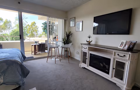 A cozy living area with a wall-mounted flat screen TV above a white wooden cabinet with a built-in electric fireplace. Two bar stools and a small table with a plant are positioned near a sliding glass door that opens to a balcony with outdoor seating and greenery visible outside. The room has carpeted flooring and light-colored walls with framed pictures.