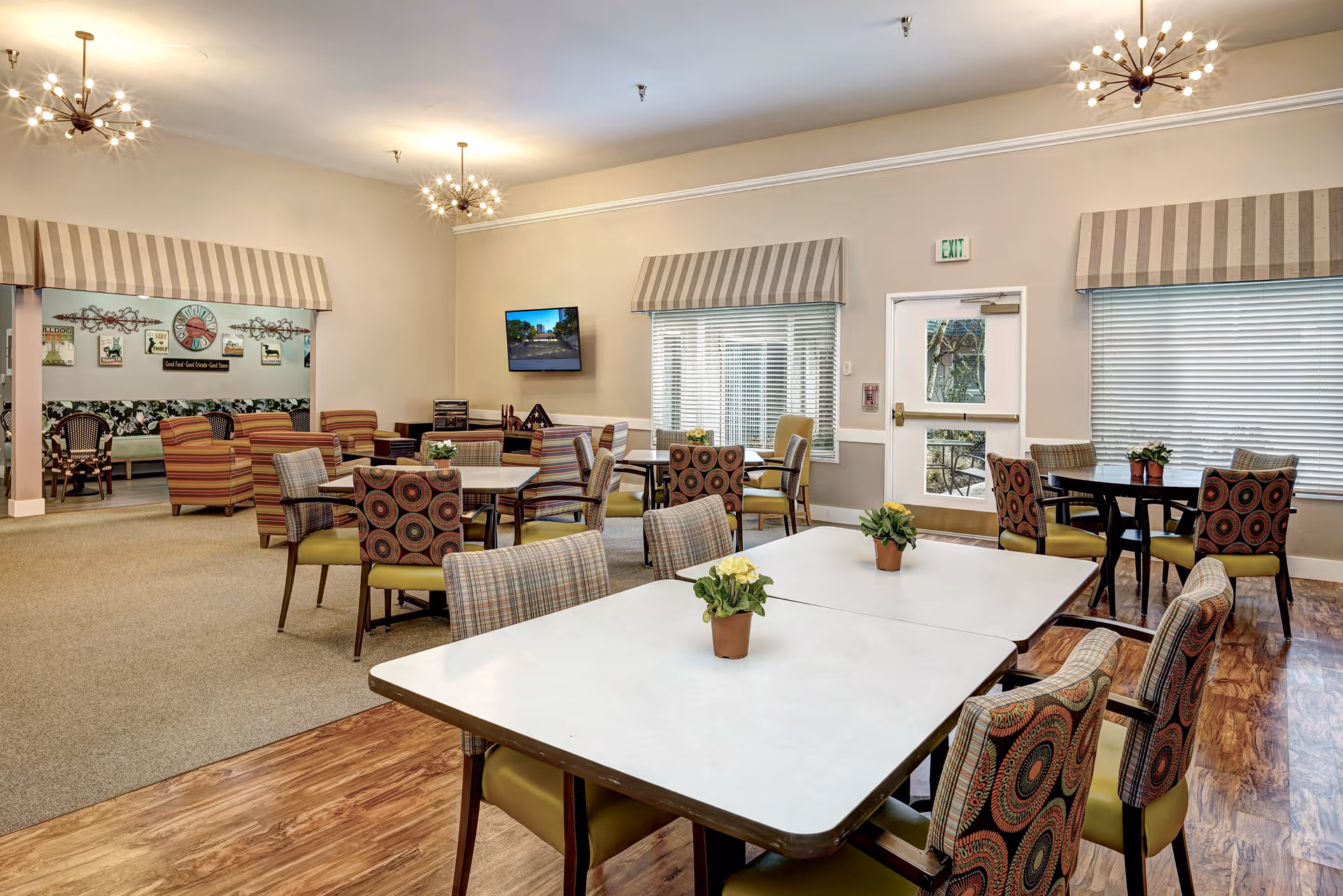 A bright and spacious common area with multiple tables and chairs arranged for dining or socializing. The room features patterned chairs, potted plants on the tables, large windows with striped valances, and modern light fixtures on the ceiling. In the background, there is a seating area with armchairs and a wall-mounted television.