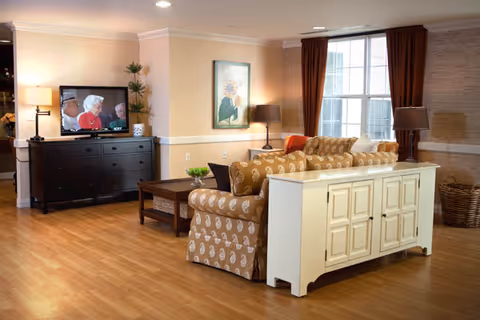A cozy living room in a senior living facility with a patterned beige sofa, a white cabinet behind the sofa, a wooden coffee table, a dark wooden dresser with a television showing elderly people, two table lamps, a framed floral painting on the wall, large windows with brown curtains, and a basket in the corner.